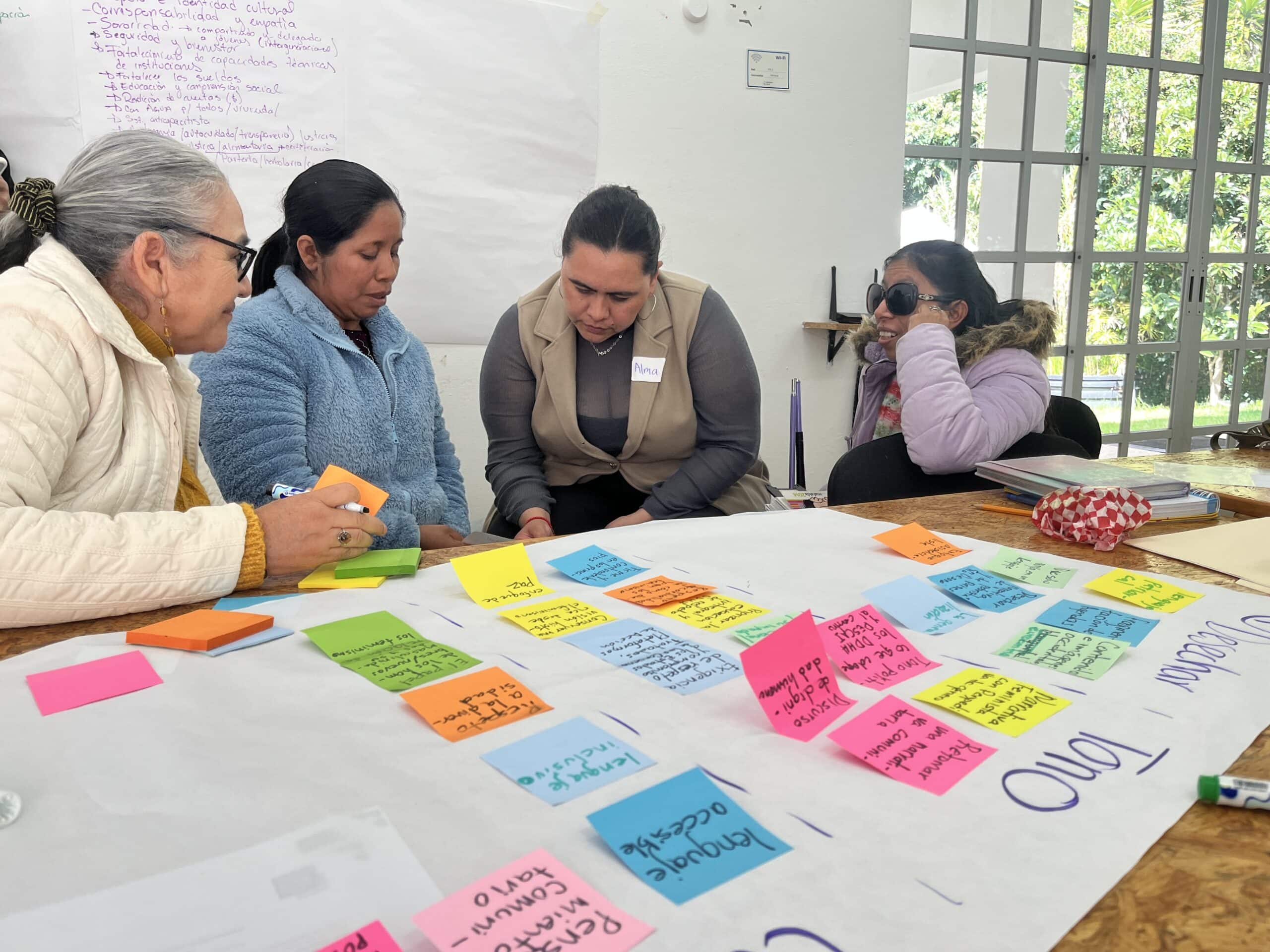 Four women sit near a large sheet of white paper covered with post-it notes. They are in discussion.